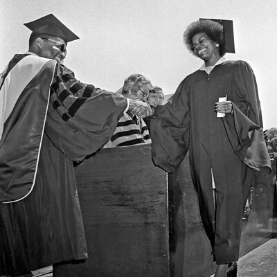 A black and white photograph from commencement. A Black woman in gown and motorboard smilingly shakes hands with a Black man dressed in doctoral robe, hood, and motorboard
