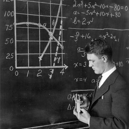 B&W photo of male student at chalkboard solving an equation