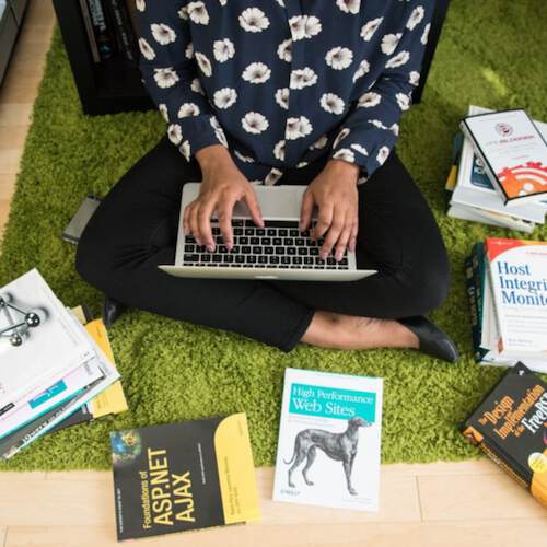 Woman sitting cross-legged on green carpet using a laptop and surrounded by books.